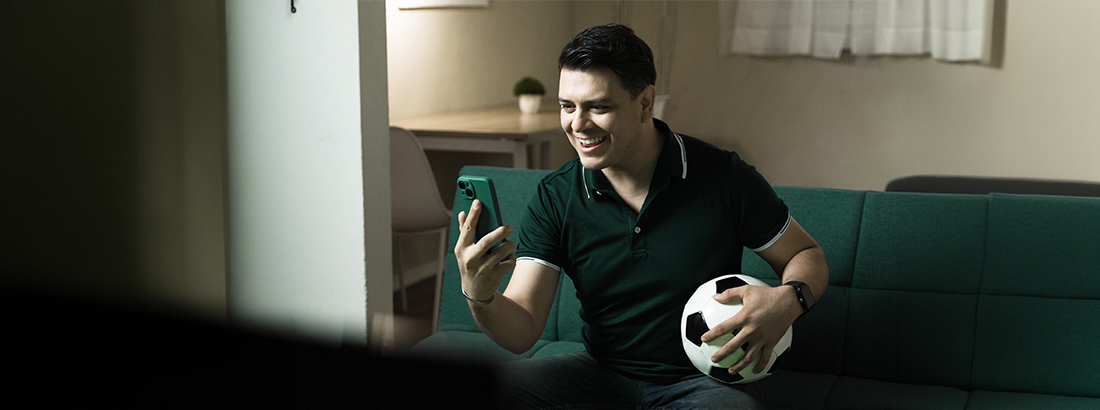 A man sitting on a couch at home, smiling while video calling on his phone and holding a soccer ball.