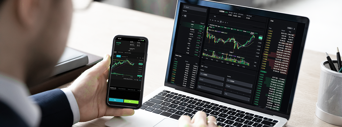 Person using a smartphone and laptop to monitor financial trading charts.